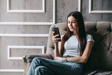 Beautiful young smiling caucasian woman resting on a sofa and using smartphone. Chatting with friends, browsing information. High quality photoの写真素材