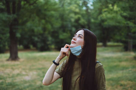 Young caucasian pretty woman removes a medical protective mask from her face on nature, surrounded...