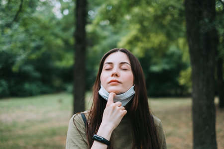 Young caucasian pretty woman removes a medical protective mask from her face on nature, surrounded by trees, breathing clean fresh air. High quality photoの写真素材