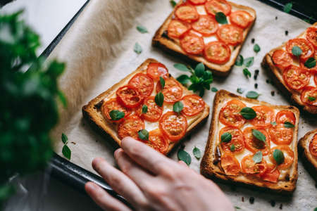 Hand takes baked sandwich with cheese and cherry tomatoes on dark bread decorated with fresh basil leaves. High quality photoの写真素材