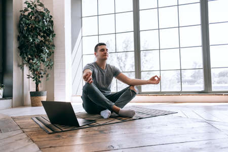 Pleased caucasian relaxed student sits crossed legs on floor near opened laptop, tries to have break after work. Yoga and meditation concept. High quality photoの写真素材
