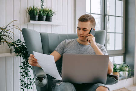 Focused young caucasian man holding paper documents sitting on a chair with laptop, calling to financial advisor or bank call center support, trouble solving.の写真素材