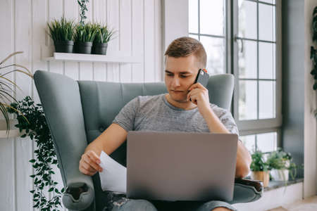 Focused young caucasian man holding paper documents sitting on a chair with laptop, calling to financial advisor or bank call center support, trouble solving.の写真素材