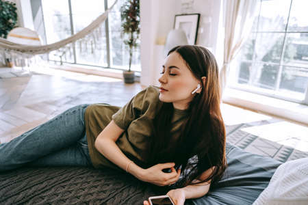 Young caucasian woman relaxing on bed at home, listening to music on headphones using smartphone on sunny day.の写真素材