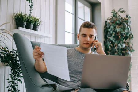 Focused young caucasian man holding paper documents sitting on a chair with laptop, calling to...