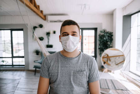 Portrait of Young brunette white European man at home on quarantine with medical mask in living room.の写真素材