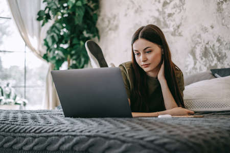 Attractive young caucasian woman working with laptop computer, lying on a bed at home. People work from home. High quality photoの写真素材