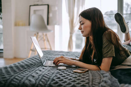 Attractive young caucasian woman working with laptop computer, lying on a bed at home. People work from home. High quality photoの写真素材