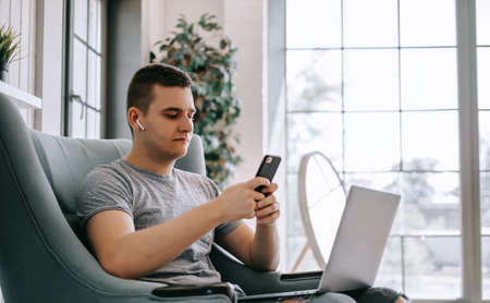 A young businessman in headphones uses a smartphone and works on a laptop, browses the internet while sitting in a chair at the home office.の写真素材