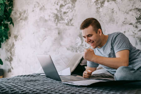 Happy young caucasian man pays bills on his laptop in the living room while lying on the bed. High quality photoの写真素材
