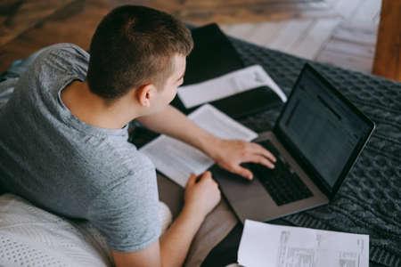 Focused young caucasian man holding paper documents on a bed with laptop, Trouble solving. Consulting about investment in internet.の写真素材