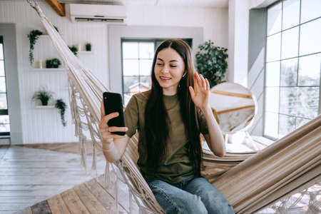 Attractive happy young woman relaxing in the home, sitting at the hammock in living room, using smartphone, speaking on video chat, waving. High quality photoの写真素材