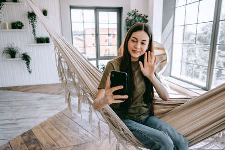 Attractive happy young woman relaxing in the home, sitting at the hammock in living room, using smartphone, speaking on video chat, waving. High quality photoの写真素材
