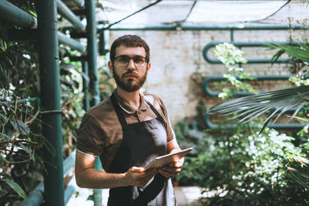 Young man gardener in glasses and apron with digital tablet working in a garden center for better...