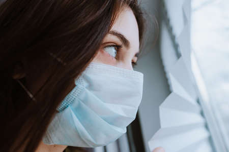 Young caucasian woman in a medical protective mask looks out the window. Coronovirus Quarantine Concept. High quality photoの写真素材
