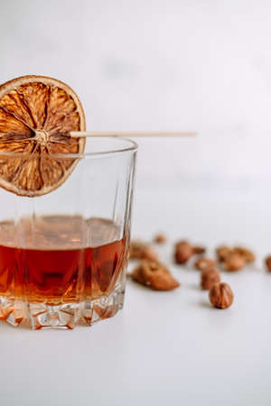 Strong alcohol whiskey in a glass cup with a slice of dried orange and nuts on a white table.の写真素材