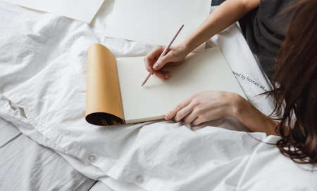 Woman artist draws in a sketchbook at home in the white bed in comfort and coziness, creativity. Drawing training.の写真素材