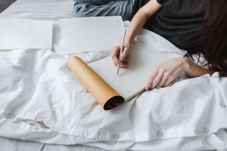 Woman artist draws in a sketchbook at home in the white bed in comfort and coziness, creativity....