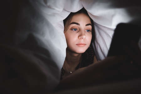 Young woman uses a smartphone, lies under a blanket on a bed at home in the evening. High quality...