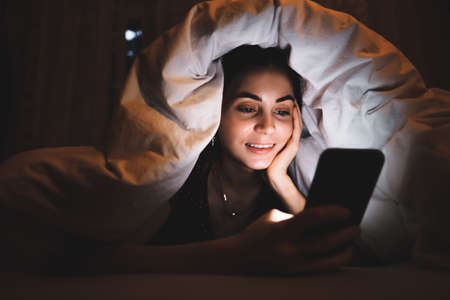 Young smiling woman uses a smartphone, lies under a blanket on a bed at home in the evening. High...