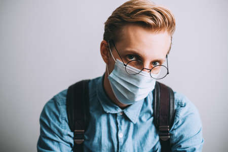 Portrait of serious young man, college student with backpack wears transparent glasses and medical disposable mask, cares about health, protects in dangerous situation.の写真素材