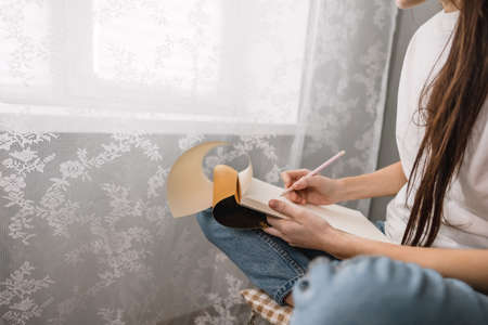 Woman artist draws in a sketchbook at home near the window, sitting on a chair in comfort and...
