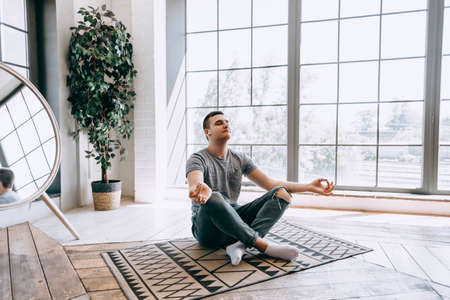 Pleased caucasian relaxed man in headphones sits crossed legs on floor near big window, tries to have break after work. Yoga and meditation concept. High quality photoの写真素材