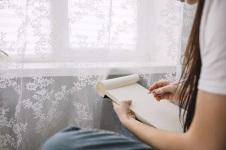 Woman artist draws in a sketchbook at home near the window, sitting on a chair in comfort and coziness, creativity. Drawing training. High quality photoの写真素材