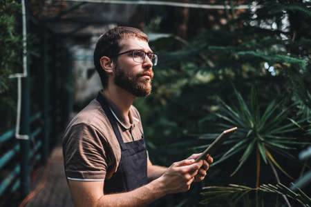 Young man gardener in glasses and apron with digital tablet working in a garden center for better...