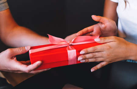 Close up of young man giving his girlfriend red gift box with pink ribbon. Woman receiving unexpected present. Celebrating concept. High quality photoの写真素材