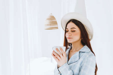 Beautiful young stylish woman in business suit and white hat standing near bed and drink coffee in sunny morning. High quality photoの写真素材