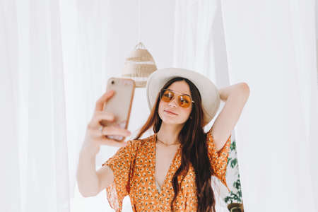 Portrait of beautiful young woman in glasses and hat while making selfie and standing near big white bed. High quality photoの写真素材