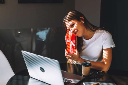 Happy young woman opening gift in front of laptop during video call or chat, celebrating birthday online. Concept of distance relations, celebrations and lifestyle. High quality photoの写真素材