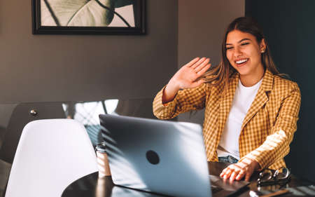 Portrait of happy beautiful stylish brunette young woman sitting at table, looking at laptop screen on video call and greeting with waving. Indoor studio shot, cafe, office background.の写真素材