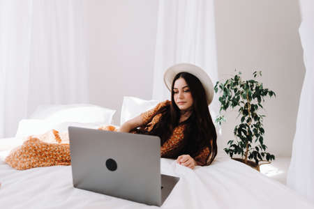 Beautiful young stylish woman in white hat lying on bed and using laptop computer in sunny morning at home. High quality photoの写真素材