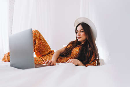 Beautiful young stylish woman in white hat lying on bed and using laptop computer in sunny morning at home. High quality photoの写真素材