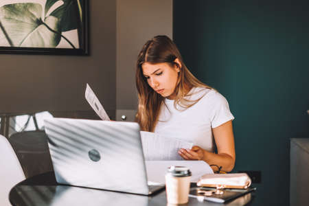 Attractive concentrated young caucasian woman calculating bills in kitchen, sitting at table with...