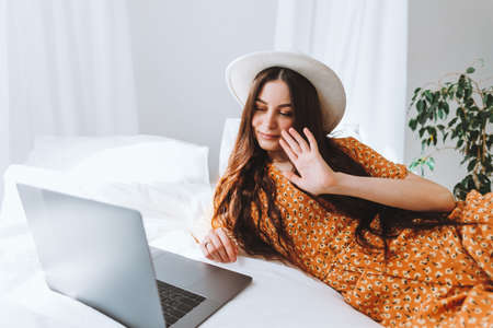 Beautiful young stylish woman in white hat lying on bed and using video call on laptop computer, in sunny morning on vacation. High quality photoの写真素材