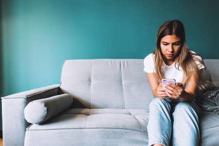 Beautiful young woman using smartphone while relaxing on sofa at living room. ... High quality photoの写真素材
