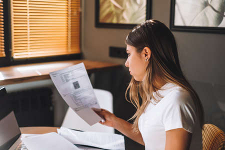 Attractive concentrated young caucasian woman calculating bills in kitchen, sitting at table with...