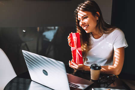 Happy young woman opening gift in front of laptop during video call or chat, celebrating birthday online. Concept of distance relations, celebrations and lifestyle. High quality photoの写真素材