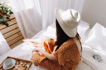 Beautiful young woman in white hat sitting on bed near window in sunny morning. Relaxing time. High quality photoの写真素材