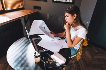 Attractive concentrated young caucasian woman calculating bills in kitchen, sitting at table with...