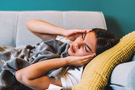 Tired young female touching forehead having headache migraine or fever, lying on a sofa in living room. ... High quality photoの写真素材