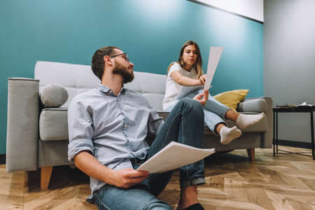 A young couple manages finances by looking through their bank accounts in the living room by the sofa. Woman and man look at documents together. Planning budget expenses. High quality photoの写真素材
