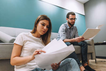 A young couple manages finances by looking through their bank accounts in the living room by the sofa. Woman and man look at documents together. Planning budget expenses. High quality photoの写真素材