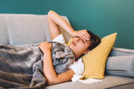 Tired young female touching forehead having headache migraine or fever, lying on a sofa in living...