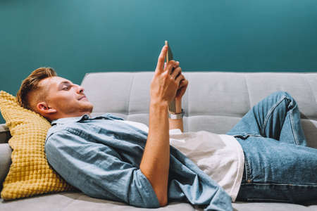 Young caucasian man lying on a sofa at home and using tablet computer, home leisure. High quality photoの写真素材
