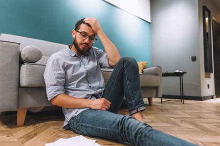 Young confused man analyzing finances at home, holding his head with hand and looking at documents. Sitting on floor near sofa in living room. High quality photoの写真素材