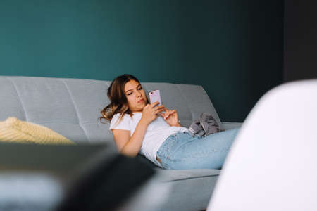 Beautiful young woman using smartphone while relaxing on sofa at living room. High quality photo